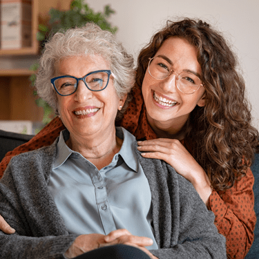 Smiling elderly woman and caregiver sitting together indoors, both wearing glasses