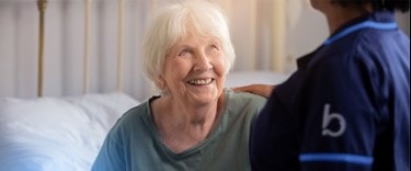 Smiling elderly woman in bed receiving comforting support from a caregiver in a navy uniform.