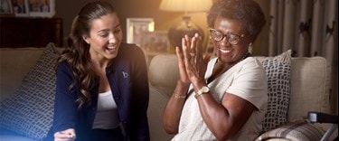 Smiling elderly woman clapping her hands while a caregiver laughs beside her on a cozy couch.