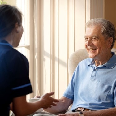 Elderly man smiling while sitting near a sunny window, talking to a caregiver in a navy uniform.