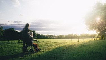A caregiver pushes an elderly woman in a wheelchair across a grassy field at sunset.