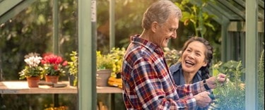 A man and woman smile joyfully together inside a vibrant greenhouse filled with lush plants.