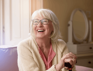 Happy older woman with glasses holding a walking cane in a bright, comfortable home setting.