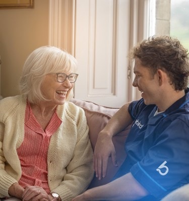 A female Bluebird Care customer sits on her sofa chatting to a Care Professional next to her