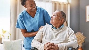 Caregiver in blue scrubs smiling warmly at an elderly man sitting in a wheelchair at home.