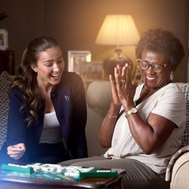 Smiling elderly woman clapping her hands while a caregiver laughs beside her on a cozy couch.