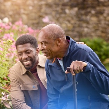 Elderly man with a walking cane and a younger man smiling together in a garden with pink flowers