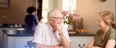 Father and daughter laughing at the kitchen table with a Bluebird Care worker in the background.