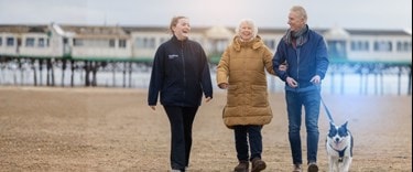 a Carer and an elederly couple walking on the beach with their dog