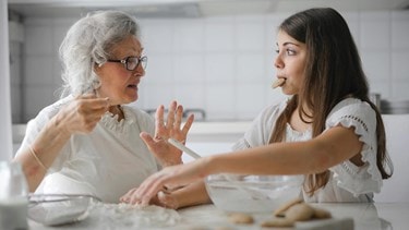Elderly woman talking while a young woman playfully eats a cookie in the kitchen during baking.