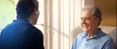 Elderly man smiling while sitting near a sunny window, talking to a caregiver in a navy uniform.