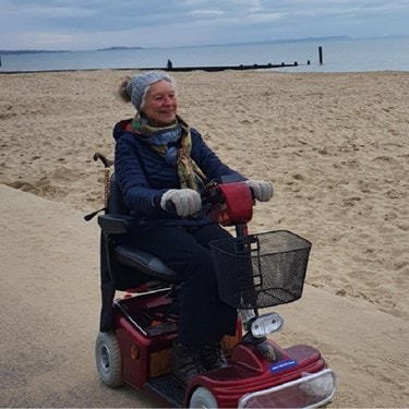 Smiling elderly woman riding a mobility scooter along a sandy beach path on a cloudy day.