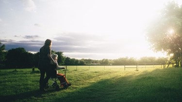 A caregiver pushes an elderly woman in a wheelchair across a grassy field at sunset.