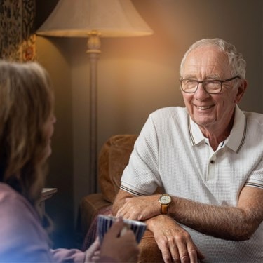 Smiling elderly man wearing glasses and a white polo shirt during a conversation at home.