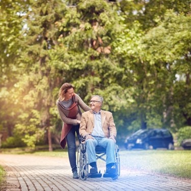 Smiling elderly man in a wheelchair being pushed by a caregiver on a scenic outdoor path