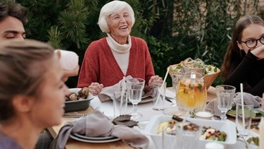 Smiling elderly woman enjoying a meal outdoors with family or friends around a table.