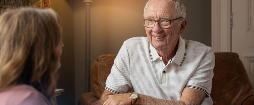 Smiling elderly man wearing glasses and a white polo shirt during a conversation at home.