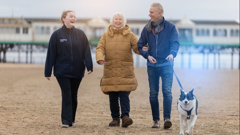 A carer walking on the beach with an elderly couple and their dog