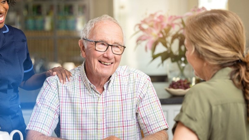 Elderly man chatting with a family member with a carer smiling