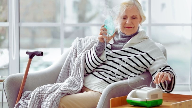 Older woman using a nebuliser at home while seated comfortably in an armchair.