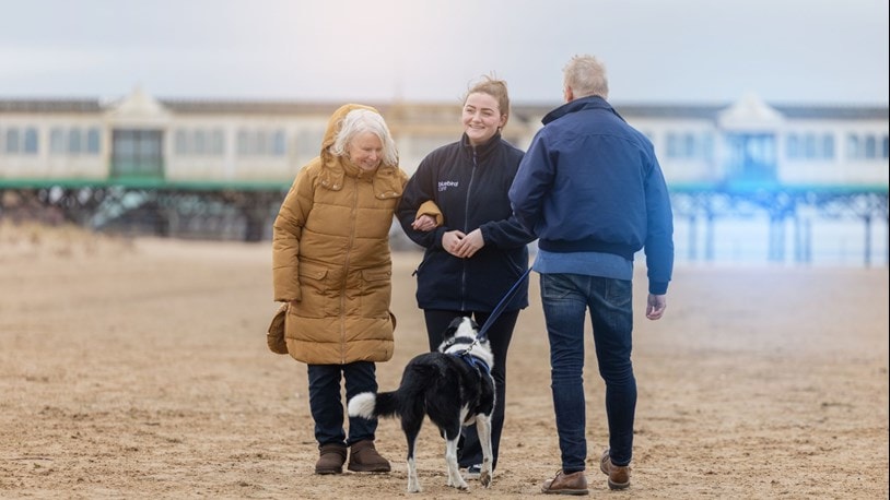 Carer walking on the beach with an elderly couple and their dog